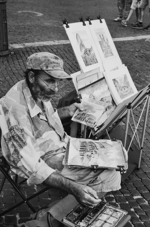 a man with hat sitting on a folding chair holding an easel and drawing some pictures