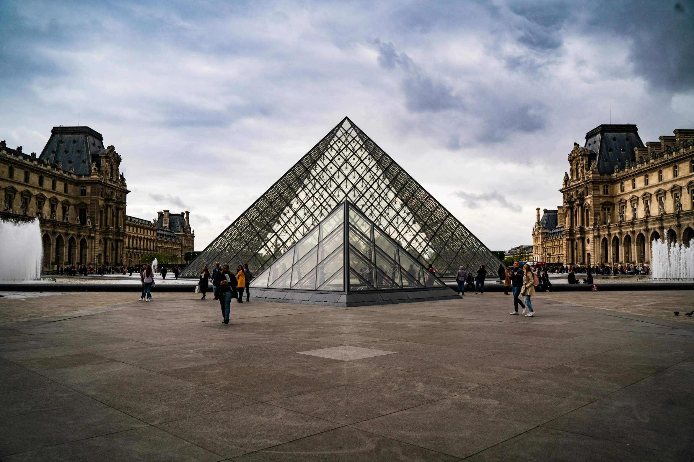 people walking around an empty cement square in front of a large pyramid