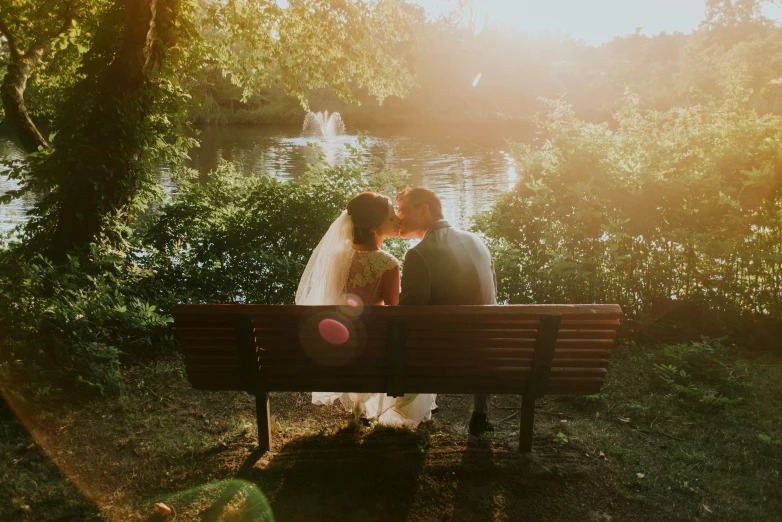 a bride and groom are kissing on a park bench