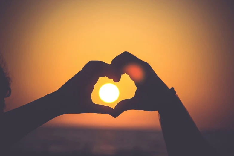 a hand making a heart shape with the sun setting behind