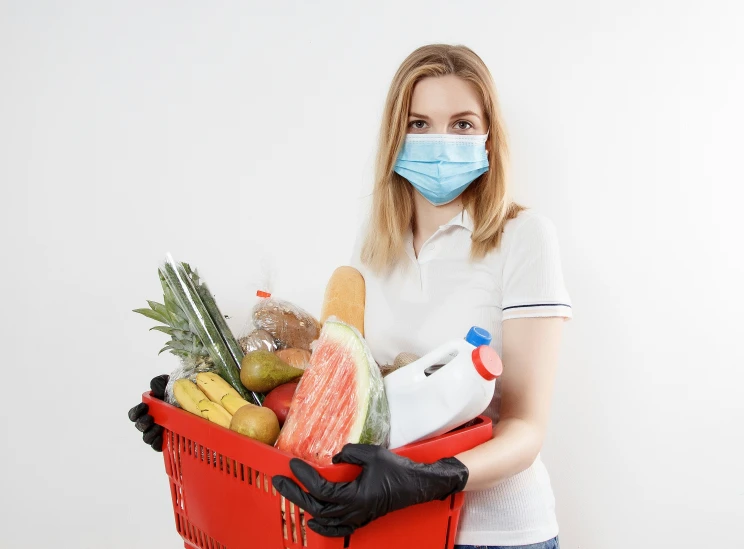 a woman wearing a mask, gloves and holding a shopping basket with food