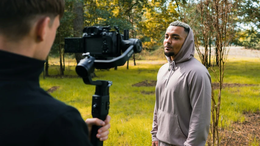 man with hood on standing in grass while holding up video camera