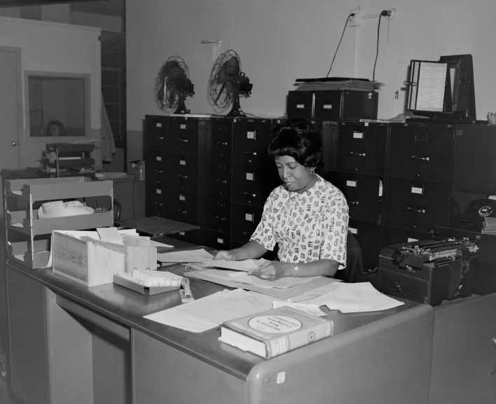 a woman sitting at a work desk in an office with files and stacks of boxes
