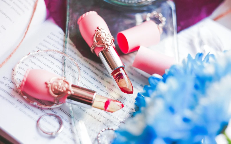 three pink lipsticks sitting on a piece of paper next to a vase with blue flowers