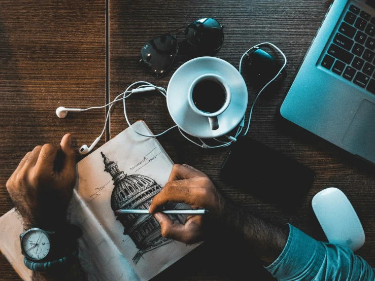 a person writing in a notebook while a cup of coffee and a laptop are on the table