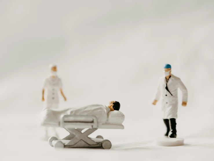 two figurines are posed by a plastic person lying on a hospital bed