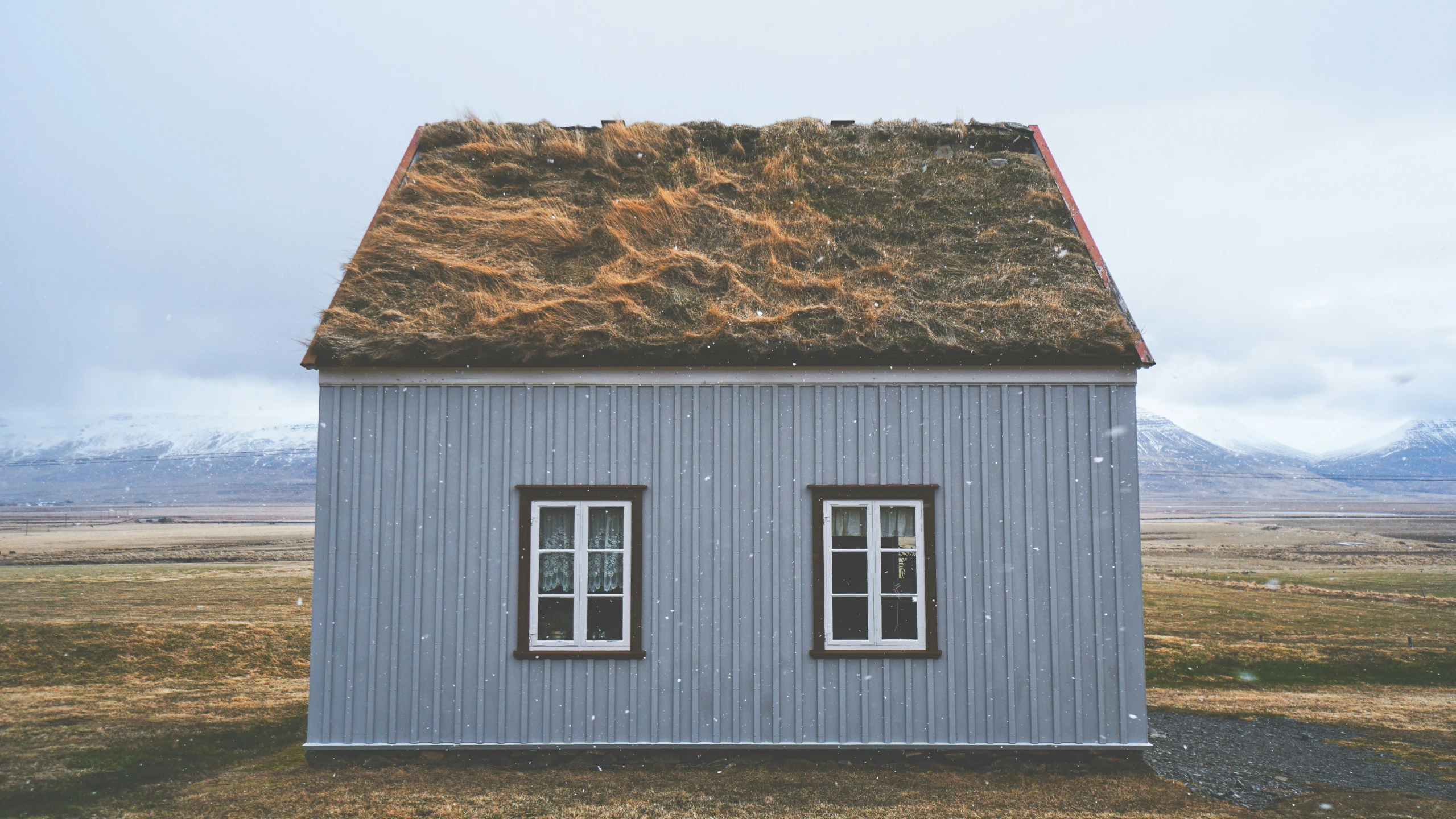 a gray cottage with three windows sits in an open field