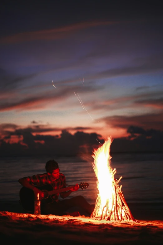 a man playing the guitar sitting by an open fire