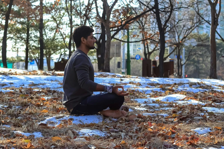 a man in a gray sweater sitting in the snow with trees and bench behind him