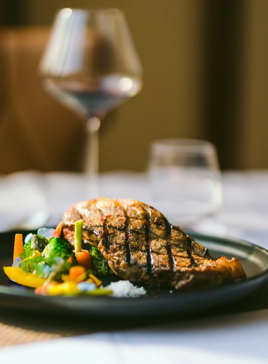 a grilled meat and vegetables on a plate with wine in the background