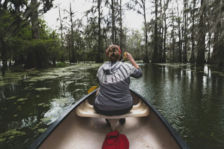 the back view of a woman in a gray jacket rowing down a river on a long canoe