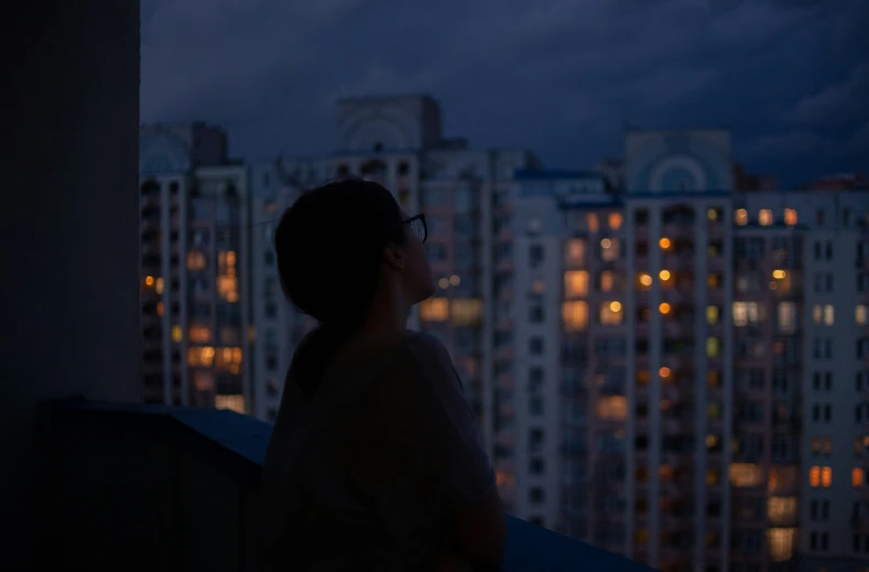 a woman on a rooftop watches out at the city skyline at night