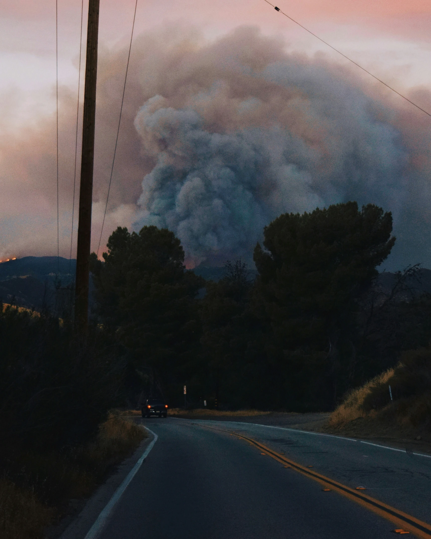 black smoke billows from an overcast sky as a car sits on the side of the road