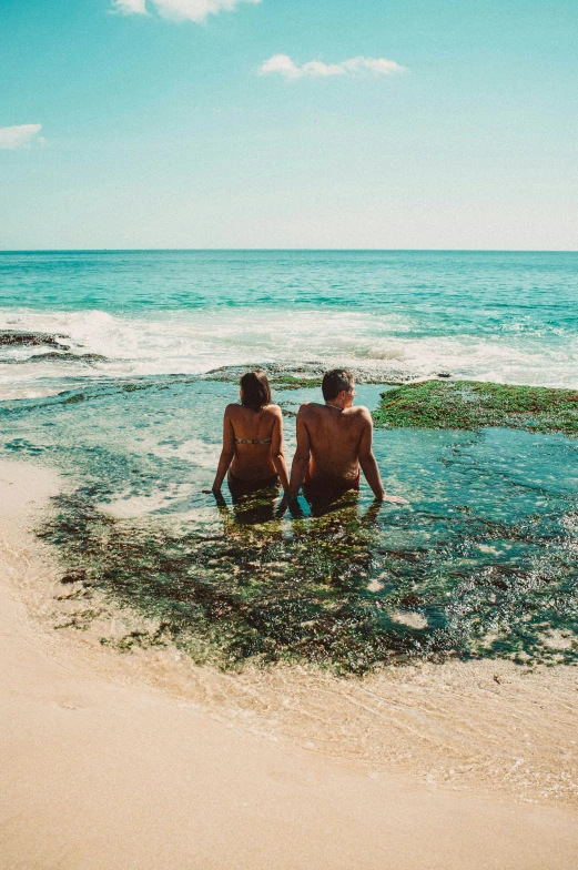 two people sitting on the beach, one facing the water