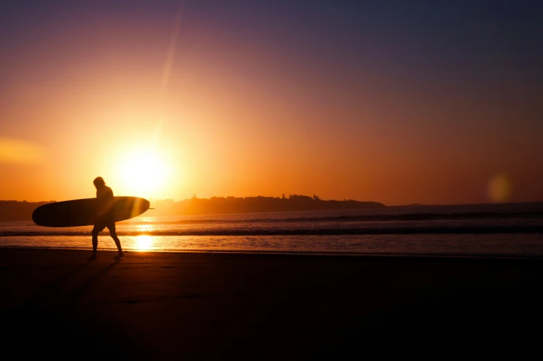 a surfer with his surfboard walks along the beach at sunrise