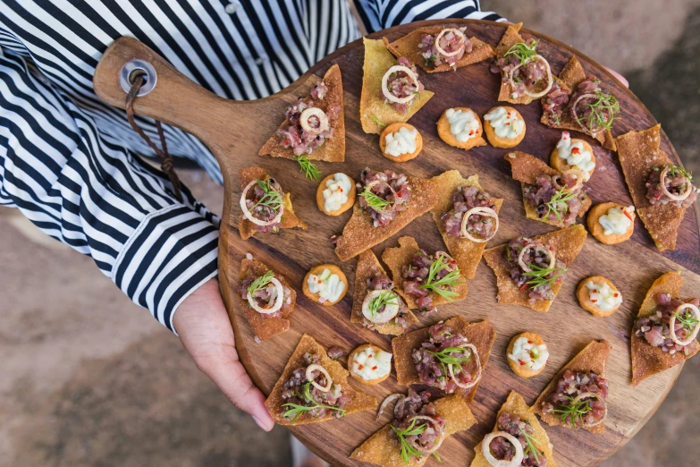 a person holding a wooden plate with small sandwiches on it