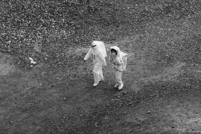 two astronauts walk in an aerial field near a rocky hill