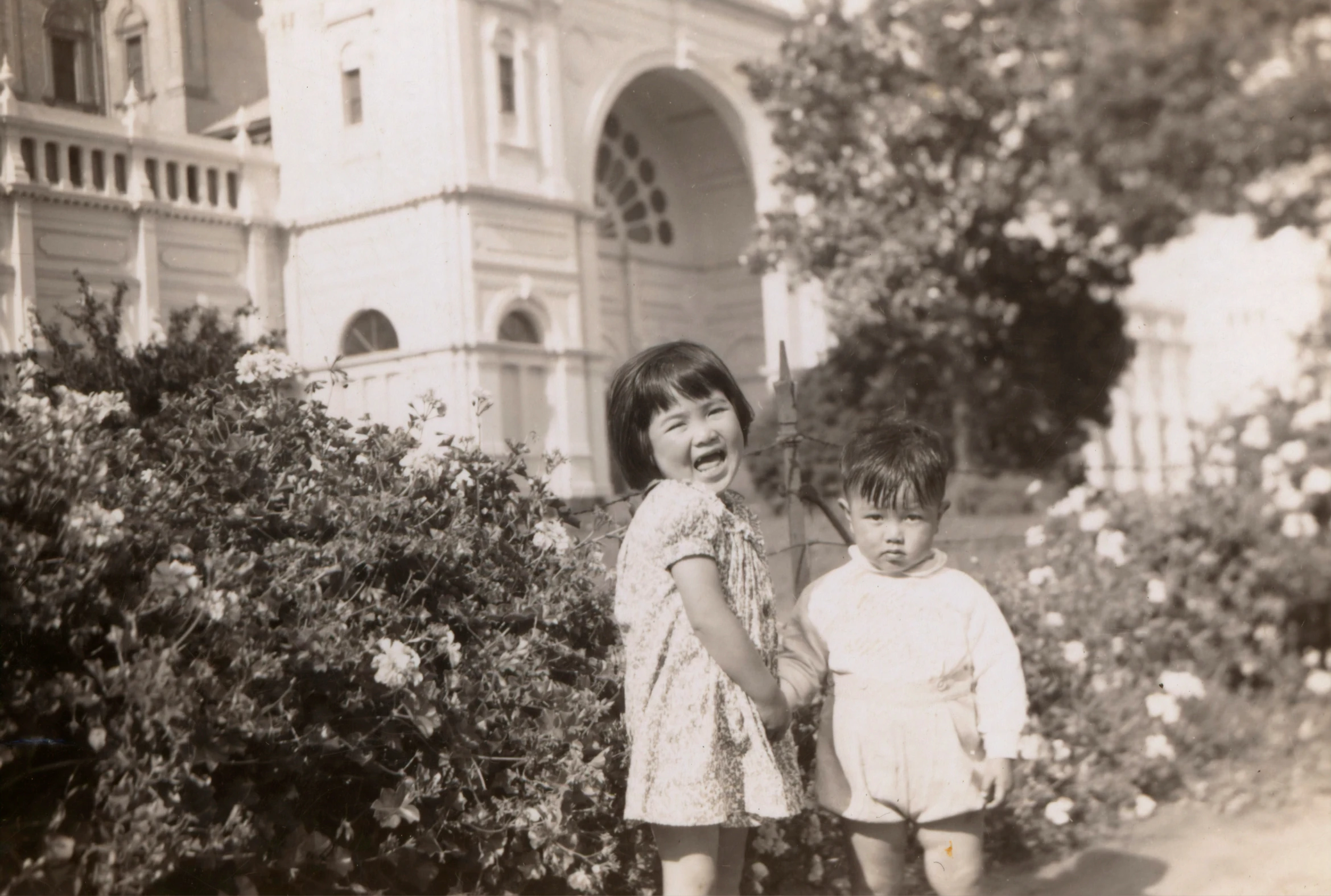 two children standing outside an old house near bushes