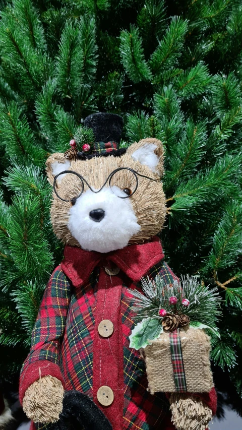 a bear ornament wearing a hat, jacket and carrying christmas presents in front of some trees