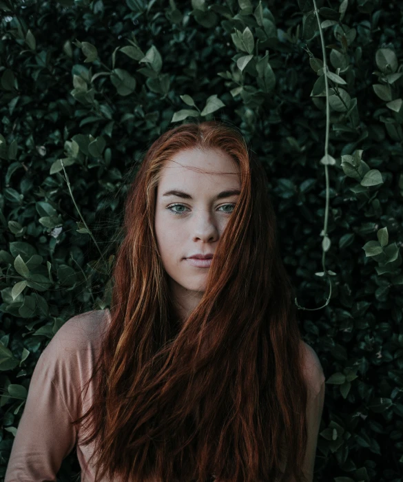 a girl with long red hair stands in front of bushes