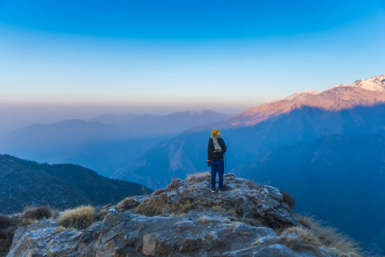 a lone hiker on the side of a mountain looks across the valley to some snowy peaks