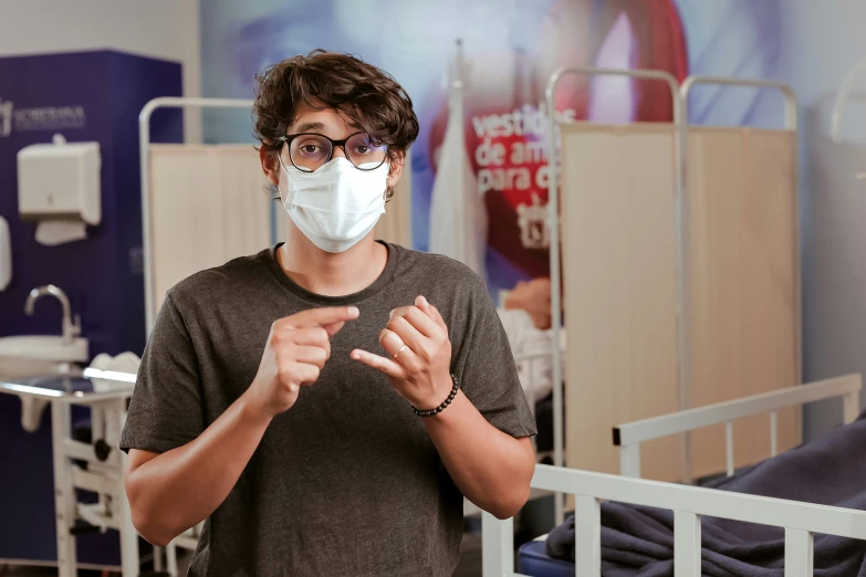 young man in grey t - shirt and black glasses making a gesture while wearing a surgical mask