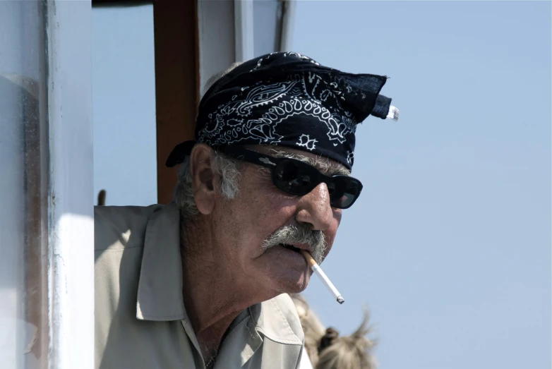 an old man with sunglasses, a bandana and a hat smokes a cigarette