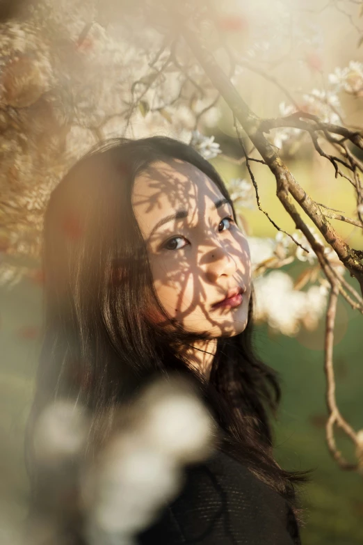 the girl with long hair stares ahead while standing under a tree