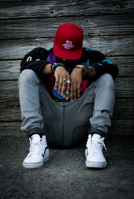 a person sitting in the shade of a wooden wall wearing white sneakers and a red hat