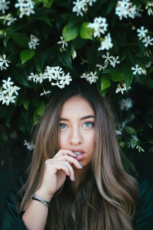a girl with her hand to her mouth, standing between flowers and trees