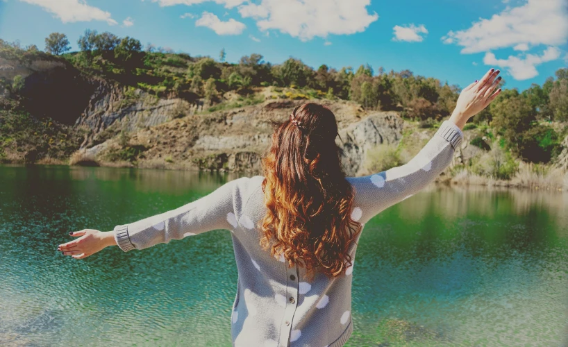 a girl standing near a body of water and waving to the sky