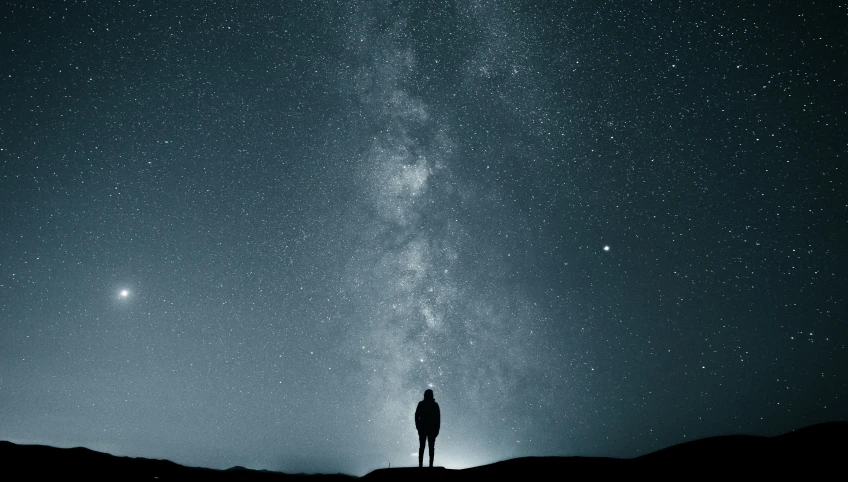 a silhouetted person standing on a hill looking up at the stars