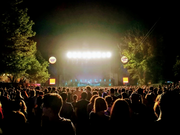 an outdoor concert with two large screens, trees and people