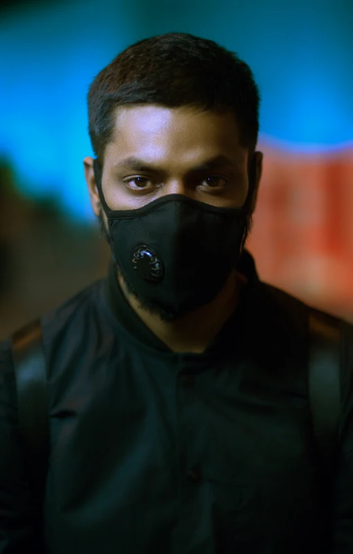 a man wearing a black mask and jacket stands in front of a brightly lit background