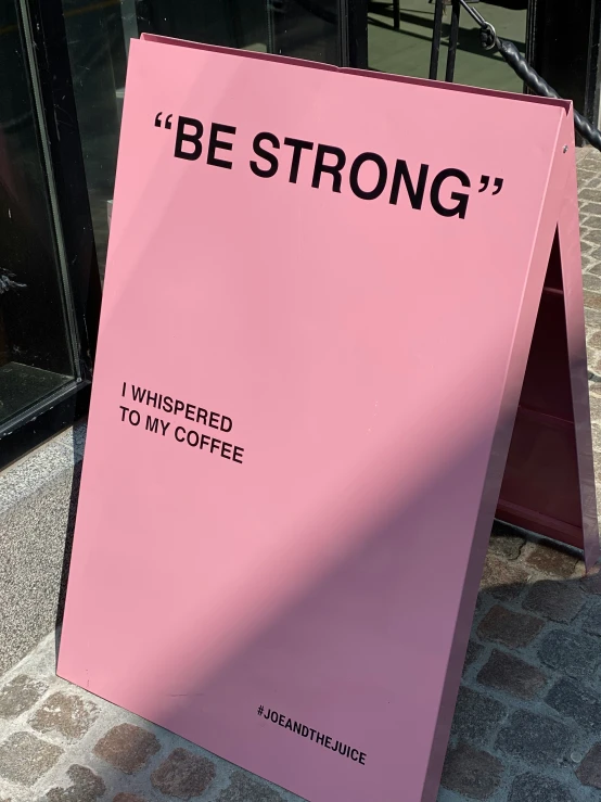 a pink sign that reads be strong, which is posted in front of a sidewalk