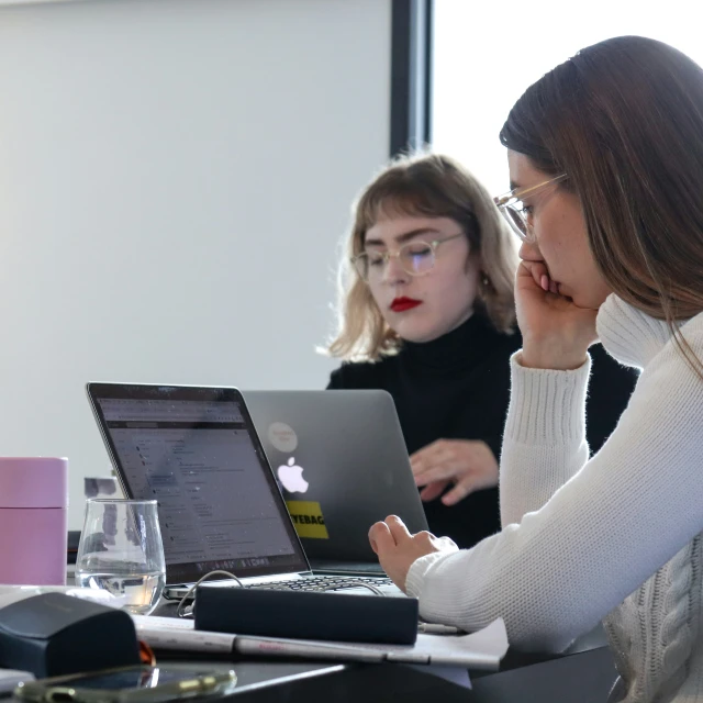 two women sitting at a table looking at an apple laptop