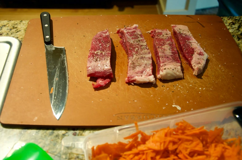 three raw pieces of meat sit on top of a chopping board near some shredded carrots