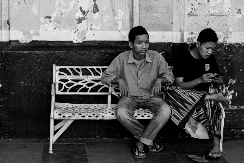 two men sit on a bench looking at their cell phones