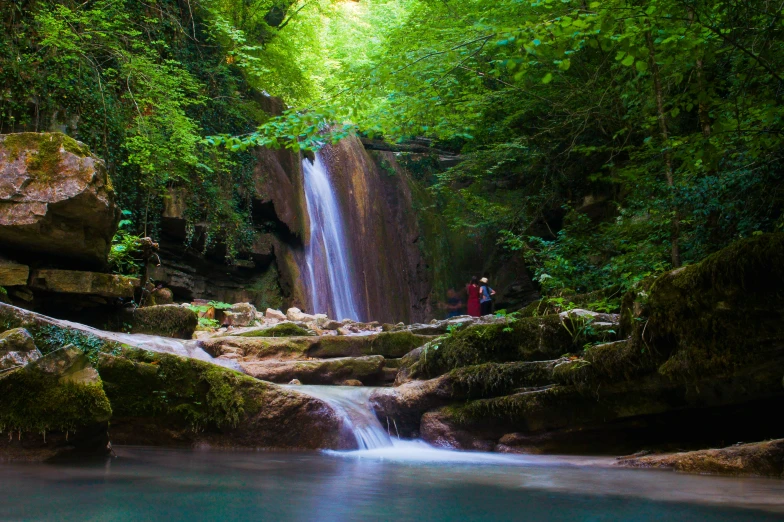 a waterfall running through a forest with two people walking