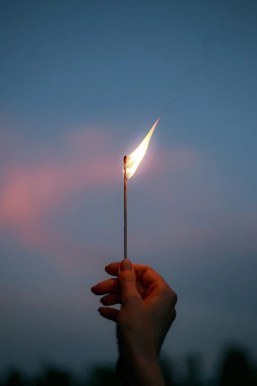hand holding lit matchstick in front of blue cloudy sky