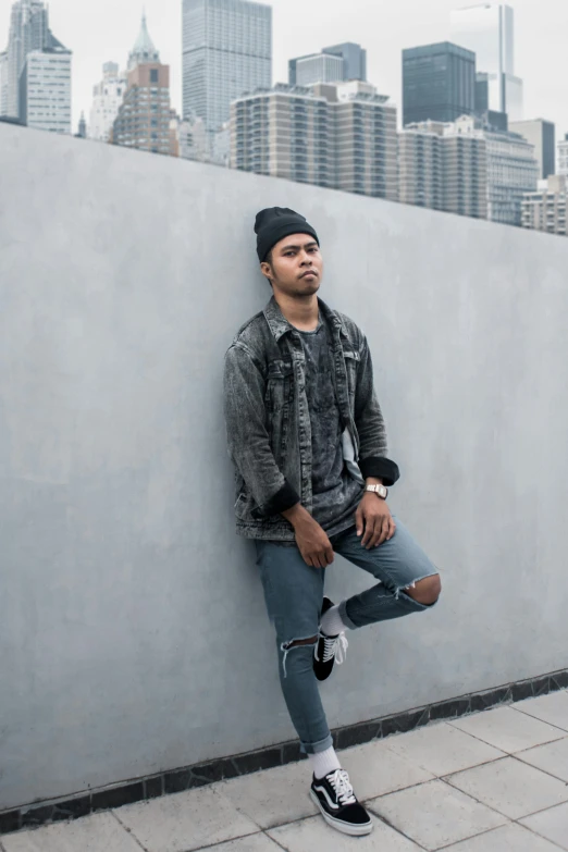 a man sitting against a cement wall in a black hat and jacket