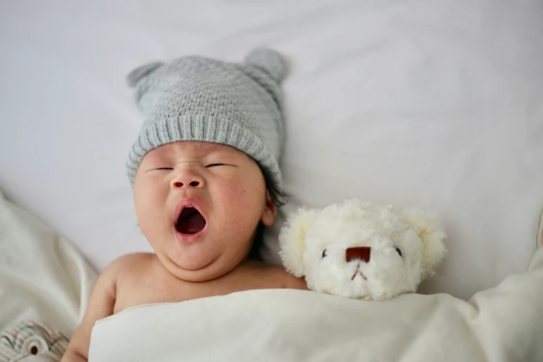 a newborn baby yawning while laying next to a stuffed bear