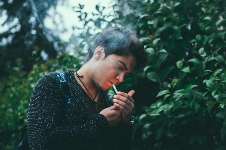 a young man smoking a cigarette with trees in the background