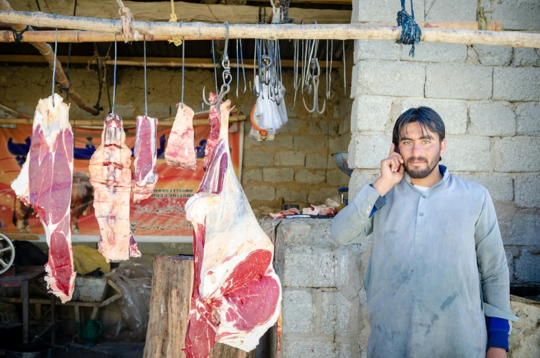 a man talking on a cell phone and standing next to different meat