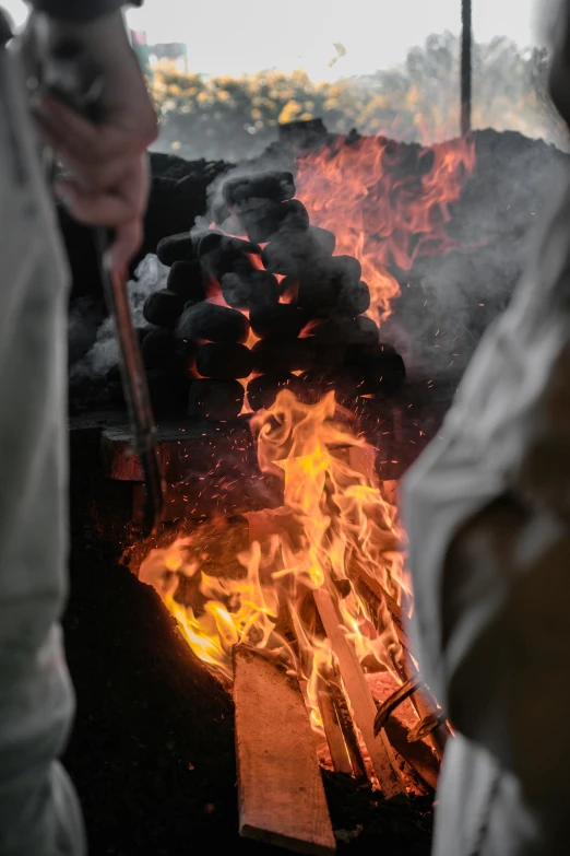 a pile of charcoalwood on fire with two people walking by