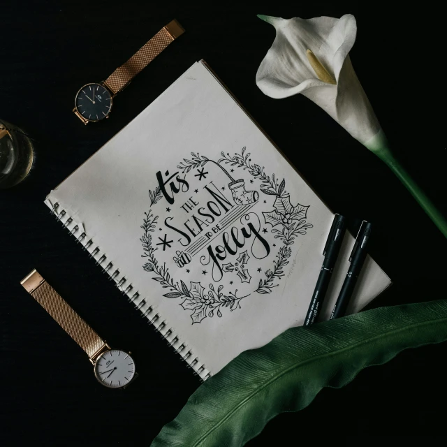 a notebook sits on a table next to a watch, some pens, and a flower