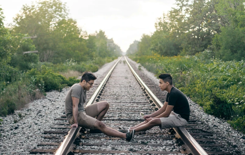 two men sitting on the train tracks touching each other