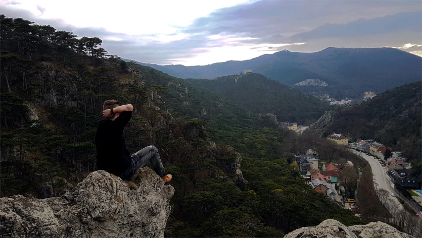 man is sitting on the edge of cliff with mountains and buildings in the background