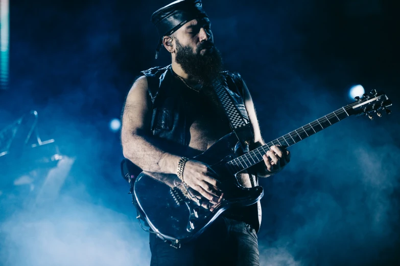a bearded man with glasses and black leather on holds an electric guitar in front of him