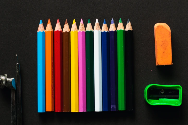 a rainbow colored pencil lined up in half and in a line next to some other pencils, sharpener and a green rubber thumb rest